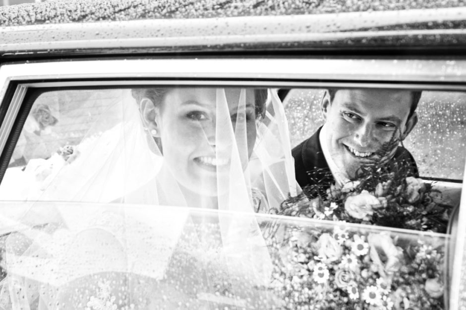 Zwart wit portret van bruidspaar in een auto op weg naar kasteel groot buggenum te Grathem voor hun trouwen.